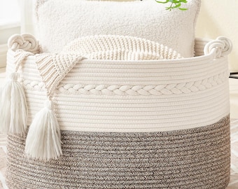May include: A large, round woven storage basket with a two-tone design. The bottom half is a dark brown, and the top half is off-white. The basket contains a white pillow, a knitted blanket, and a decorative tassel.