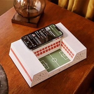 May include: A white and red stadium-shaped phone stand with a green football pitch design inside. A smartphone displays a music app. The stand has speaker holes and a red stripe. The phone screen reads "The Best in Football".