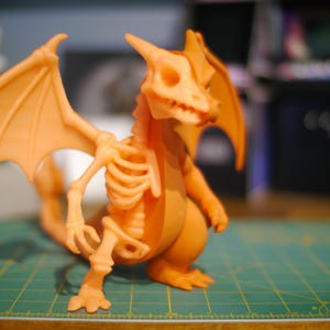 Puede incluir: Figura de dragón naranja impresa en 3D con diseño esquelético. El dragón tiene alas grandes, una cola larga y una caja torácica detallada. La figura está colocada sobre una esterilla de corte verde.