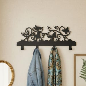 Könnte beinhalten: Eine schwarze Wandgarderobe aus Metall mit einem dekorativen oberen Rand, der Schmetterlings- und Rankenmotive aufweist. Die Garderobe hat vier nach unten gerichtete Haken, an denen eine blaue Jeansjacke und ein gemusterter türkiser Schal hängen.