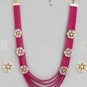 Könnte beinhalten: Eine magenta Perlenkette mit passenden Ohrringen. Die Halskette hat mehrere Stränge aus kleinen Perlen und ist mit blütenförmigen Anhängern mit roten und weißen Akzenten verziert. Die Ohrringe haben ein ähnliches Design.