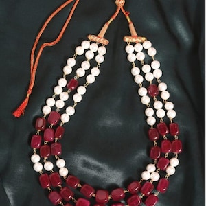 May include: A three-strand necklace with alternating rows of white, round beads and rectangular, deep red beads. The necklace has a gold-coloured chain and an orange cord with a tassel for adjusting the length. The background is a dark green fabric.