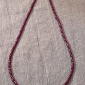 Könnte beinhalten: Eine Halskette aus kleinen, facettierten, tiefrosa Edelsteinen. Die Perlen sind zu einer durchgehenden Schlaufe aufgereiht. Der Verschluss ist silberfarben. Die Halskette wird auf einer neutralen, strukturierten Oberfläche präsentiert.