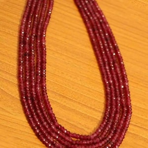 Op de afbeelding: Een ketting met meerdere strengen met facetgeslepen, dieprode kralen. De ketting heeft een symmetrisch ontwerp, waarbij de strengen naar beneden vallen. De kralen zijn aan elkaar geregen, waardoor een vloeiende, elegante uitstraling ontstaat. De ketting wordt gepresenteerd op een houten oppervlak.