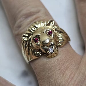 May include: A gold-colored lion ring with ruby eyes and a diamond in its mouth. The ring is highly detailed, with a textured mane and facial features. The ring is worn on a finger.