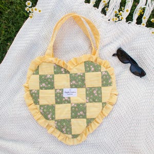May include: Heart-shaped patchwork tote bag with yellow and green floral squares, yellow ruffle trim, and matching handles. The bag rests on a white blanket with fringe and daisies. Black sunglasses are nearby.