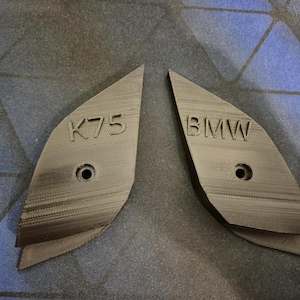 Peut inclure: Deux pièces angulaires grises avec les inscriptions "K75" et "BMW" en relief. Chaque pièce comporte un trou circulaire. Les articles semblent être imprimés en 3D, possiblement des pièces ou accessoires automobiles.