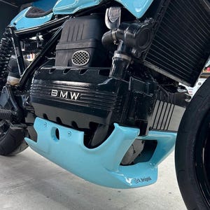Puede incluir: Primer plano de un motor de motocicleta personalizado. El motor es negro con el logotipo de BMW y detalles en azul claro en el carenado. La motocicleta tiene un neumático negro y un radiador negro.