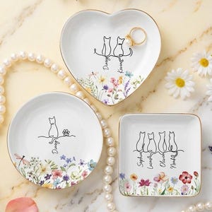 Könnte beinhalten: Drei Keramik-Schmuckschalen in Herz-, runder und quadratischer Form. Jede Schale hat ein schwarzes Katzensilhouetten-Design mit floralen Akzenten und goldenem Rand. Namen sind unter den Katzen geschrieben. Eine Perlenkette und Blumen umgeben die Schalen.
