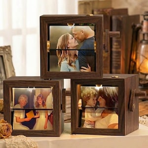 Könnte beinhalten: Drei Holz-Fotowürfel mit drehbaren Bildtafeln. Jeder Würfel zeigt ein anderes Familienfoto, das durch eine Innenbeleuchtung beleuchtet wird. Die Würfel sind dunkelbraun und haben eine Vintage-Ästhetik mit einem warmen, einladenden Glanz.