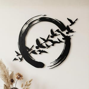 Könnte beinhalten: Schwarze Metall-Wandkunst mit einem Vogelschwarm im Flug, der aus einem großen, stilisierten schwarzen Kreis hervorgeht. Das Kunstwerk ist an einer weißen Wand befestigt und erzeugt einen auffälligen Kontrast. Die Vögel sind als Silhouette dargestellt.