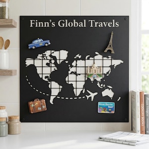 Op de afbeelding: Een zwarte metalen wereldkaart met witte uitgesneden continenten en rastervierkanten. De tekst "Finn's Global Travels" staat bovenaan. Decoratieve magneten zijn onder meer een blauwe taxi, de Eiffeltoren, de Taj Mahal, een koffer, een vliegtuig en een Great Barrier Reef-magneet.