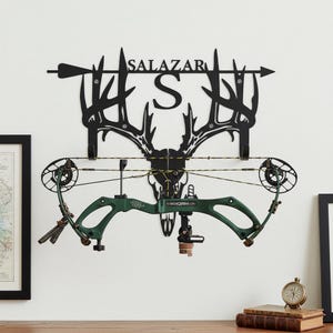 May include: Black metal wall-mounted bow rack with the name "SALAZAR" and a large letter "S". The rack features a deer skull design and holds a green compound bow. An arrow is on the left and right side of the name.