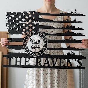 May include: Black metal wall art shaped like the American flag, featuring the US Navy emblem and silhouettes of ships. The text "THE RAVEN'S" is displayed at the bottom. The flag design includes stars and stripes, with a distressed look.