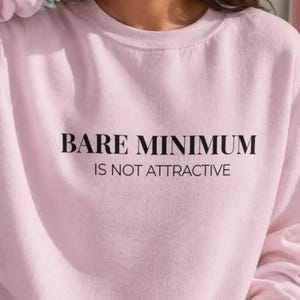 Puede incluir: Sudadera rosa claro con el texto negro "BARE MINIMUM IS NOT ATTRACTIVE". La sudadera es de cuello redondo y parece estar hecha de un material suave. La imagen es un primer plano de la sudadera.