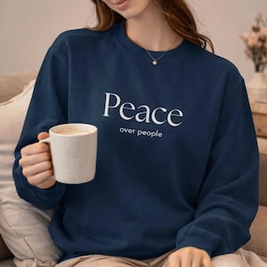 Puede incluir: Sudadera azul marino con la frase "Peace over people" en blanco. Una taza blanca se sostiene en la mano. La sudadera es de cuello redondo.