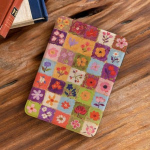May include: A notebook cover with a floral patchwork design. The cover displays a variety of colourful flowers in square blocks, including pink, orange, purple, and yellow blooms. The notebook is on a wooden surface.