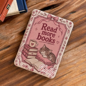 May include: A rectangular e-reader case in a dusty rose color featuring a graphic of a sleeping tabby cat on a pillow, a stack of books, a coffee mug, and the text Read more books and the name Dolly in a cursive script font.