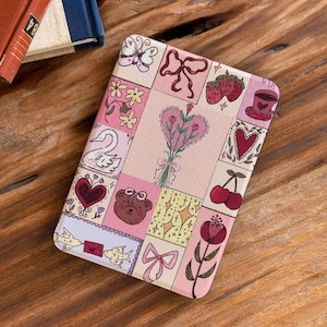 May include: A pink and cream-colored case with a patchwork design featuring hearts, flowers, bows, and a teddy bear. The case has a rectangular shape and is likely for an electronic device.
