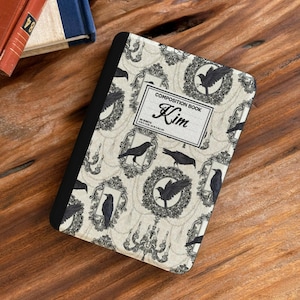 Può includere: Un quaderno di composizione rettangolare con dorso nero e copertina color crema caratterizzata da un motivo ripetuto di corvi neri, cornici ornate e candelabri. L'etichetta centrale riporta la scritta COMPOSITION BOOK e il nome Kim.