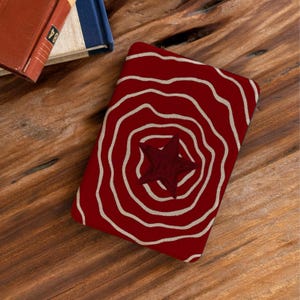 Può includere: Una copertina di libro rossa con un disegno a stella al centro, circondata da cerchi concentrici bianchi. La copertina è su una superficie di legno, con i bordi di due libri visibili sullo sfondo.