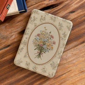 Op de afbeelding: Een rechthoekige beige boekomslag met een bloemenpatroon en een ovaalvormig centraal ontwerp. Het ontwerp toont een boeket van roze, gele en blauwe bloemen, vastgebonden met een roze lint. De omslag ligt op een houten oppervlak.