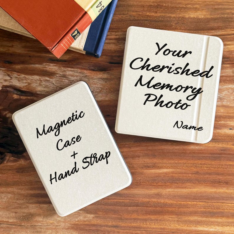 May include: Two rectangular off-white cases with black text. One case reads Your Cherished Memory Photo Name. The other case reads Magnetic Case + Hand Strap. The items are positioned on a wooden surface near stacked books.