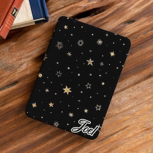 May include: A black rectangular e-reader cover featuring a pattern of gold-colored stars in various sizes and styles. The name Joel is printed in a white cursive script font in the bottom right corner of the cover, which has rounded corners.
