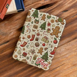May include: A rectangular e-reader cover featuring a cream-colored background with a repeating Christmas pattern of gingerbread people, green pine trees, red stockings, wrapped gift boxes, and ornaments, with the name Faye written in red script.