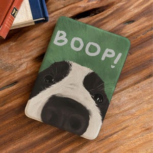 Może przedstawiać: Prostokątna zielona mata z białym napisem "BOOP!". Mata przedstawia zbliżenie twarzy czarno-białego psa, z widocznym nosem i oczami. Mata leży na drewnianej powierzchni.