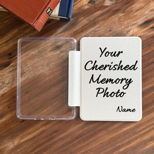 May include: A rectangular, hinged photo frame with a clear plastic left side and a solid white right side. The white panel features black cursive text that reads: Your Cherished Memory Photo, with the word Name printed below it.