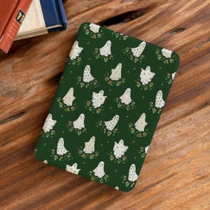 Könnte beinhalten: Eine dunkelgrüne Hülle mit einem sich wiederholenden Muster aus weißen Geistern und floralen Details. Die Geister haben verschiedene Designs und sind vor einem Hintergrund aus kleinen Blumen angeordnet. Die rechteckige Hülle ist wahrscheinlich für einen E-Reader oder ein Tablet.