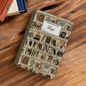 May include: A rectangular notebook with a brown spine and a cover featuring a repeating pattern of bookshelves filled with various books, clocks, and vases. A white rectangular label is centered on the cover with the text COMPOSITION BOOK Faye.