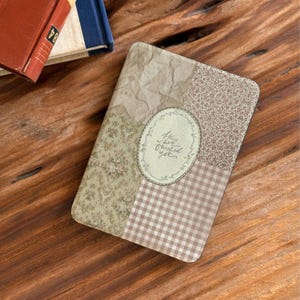 Puede incluir: Un cuaderno rectangular con un diseño de patchwork. La cubierta presenta secciones de papel floral, a cuadros y texturizado en tonos beige y marrón. Un marco ovalado en el centro contiene el texto "Life's not finished yet."