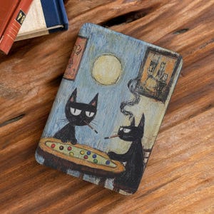 Op de afbeelding: Een rechthoekige telefoonhoes met een artistieke illustratie van twee zwarte katten die poolen. De katten roken sigaretten, met een volle maan en ingelijste kunstwerken op de achtergrond. De hoes ligt op een houten oppervlak.
