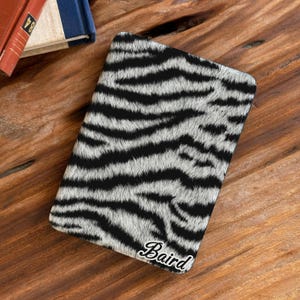 May include: A rectangular e-reader cover featuring a black and white zebra stripe pattern with a textured, fur-like appearance. The bottom right corner displays the name Baird in a white, cursive, script-style font with a black outline.