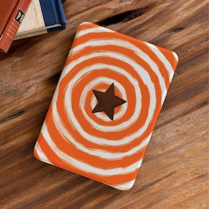 May include: A rectangular e-reader cover featuring a vibrant orange and white spiral pattern that radiates from the center. A solid dark brown five-pointed star is positioned directly in the middle of the swirling design on the front surface.