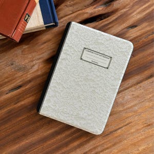 Puede incluir: Un cuaderno de composición con una cubierta de encaje blanco. El cuaderno tiene un lomo negro y las palabras "COMPOSITION NOTEBOOK" y "SALEM HILLS" impresas en la parte delantera. Otros libros son visibles en el fondo.