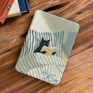 May include: A rectangular e-reader case featuring a graphic illustration of a black cat reading a book while tucked into a bed with blue and white striped bedding. The name Kim is printed in a white cursive script at the bottom of the cover.