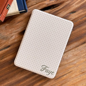 May include: A rectangular e-reader cover in a light beige color featuring a repeating pattern of small dark gray dots. The bottom right corner of the cover displays the name Faye in a cursive script font. The cover has rounded corners.