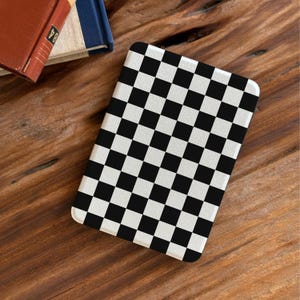 Pode incluir: Um objeto retangular com um padrão xadrez preto e branco. O objeto está sobre uma superfície de madeira, com a esquina de um livro ao fundo. O padrão é um design clássico de tabuleiro de xadrez.
