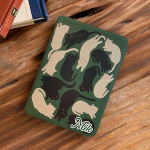 May include: A rectangular dark green case featuring a repeating pattern of illustrated black and cream-coloured cats in various resting positions. The name Milo is printed in a white cursive script font in the bottom right corner of the case.
