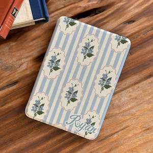 May include: A rectangular e-reader cover featuring a light blue and white vertical striped pattern with repeating scalloped white frames containing blue floral illustrations and the name Ryan written in a cursive script at the bottom right.