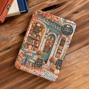 May include: A rectangular e-reader case featuring a colorful autumn illustration of a bookstore storefront named Cider & Stories. The design includes orange maple leaves, pumpkins, a black cat, and a table with chairs. The name Bobbi is printed at the bottom.