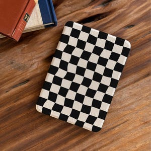 May include: A rectangular coaster with a black and white checkered pattern. The design features a wavy, distorted checkerboard effect. The coaster is set on a wooden surface, with the corner of a book visible in the background.