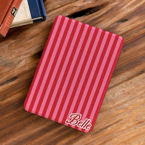 May include: A rectangular e-reader cover featuring a vertical striped pattern in alternating shades of red and light pink. The name Belle is printed in a white cursive script font with a red outline in the bottom right corner of the cover.