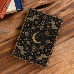 Pode incluir: Um livro retangular com capa preta e um design celestial dourado. A ilustração mostra uma lua crescente central rodeada por estrelas radiantes, padrões de sol e nuvens dispersas. A borda apresenta um padrão geométrico decorativo.