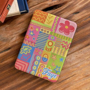 May include: A rectangular e-reader cover featuring a vibrant patchwork design of colourful geometric shapes, wavy lines, stars, and a large red flower. The name Faye is printed in a white script font at the bottom right corner of the cover.