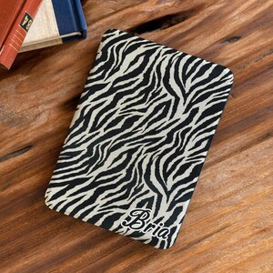 May include: A rectangular e-reader cover featuring a black and white zebra stripe pattern. The name Bria is printed in a white cursive script font near the bottom right corner of the cover. The cover has rounded corners and a smooth surface.