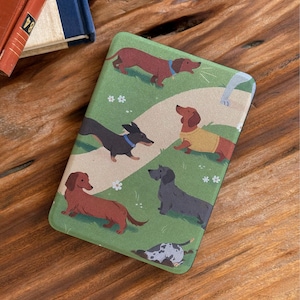 May include: A rectangular case with a playful design of dachshund dogs on a green backdrop. The dogs are shown in different poses and colours, with a path meandering through the scene. White flowers and a light blue collar add detail.
